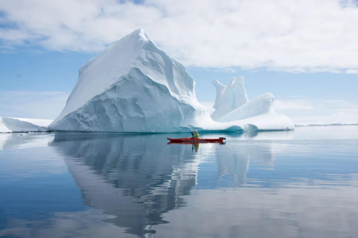 Person in red kayak paddling near large jagged iceberg in calm, reflective water.