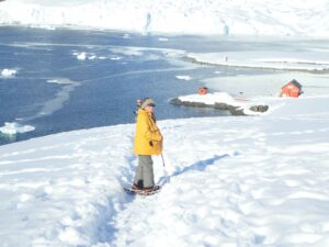 "Person wearing a yellow jacket and snowshoes stands in snowy Antarctic landscape, near ice floes and research stations.