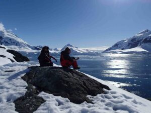 Two people sitting on a snow-covered rock by a body of water with snow-covered mountains in the background in Antarctica.