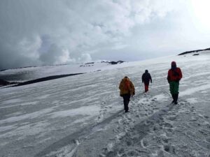 Three people walking on snowy Antarctic terrain with winter gear, cloudy sky and rugged, icy landscape.