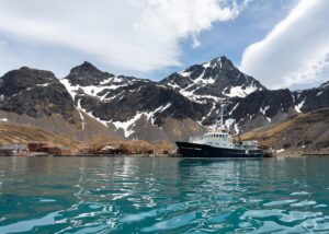 Docked ship near snow-covered mountains with patches of blue sky and clouds in Antarctica.