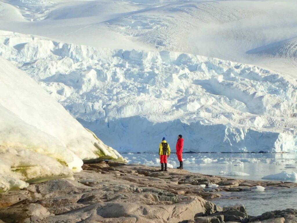 Two people standing near the edge of icy water with a glacier in the background in Antarctica.