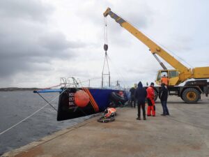 A crane lifting a boat out of the water at a dock during the Volvo Ocean Race, with crew members in red and black outfits standing around.