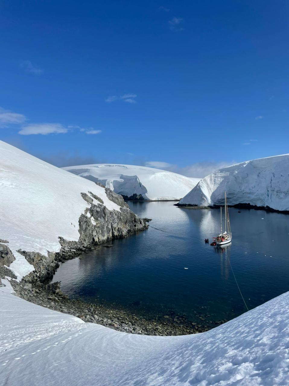 Sailboat in Antarctica's icy waters surrounded by snow-covered cliffs under a clear blue sky.