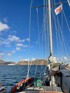 Sailboat on the water equipped with various ropes and equipment, with a flag on one of the masts. The background features a clear blue sky with clouds and a mountainous coastline, likely near Deception Island in Antarctica.