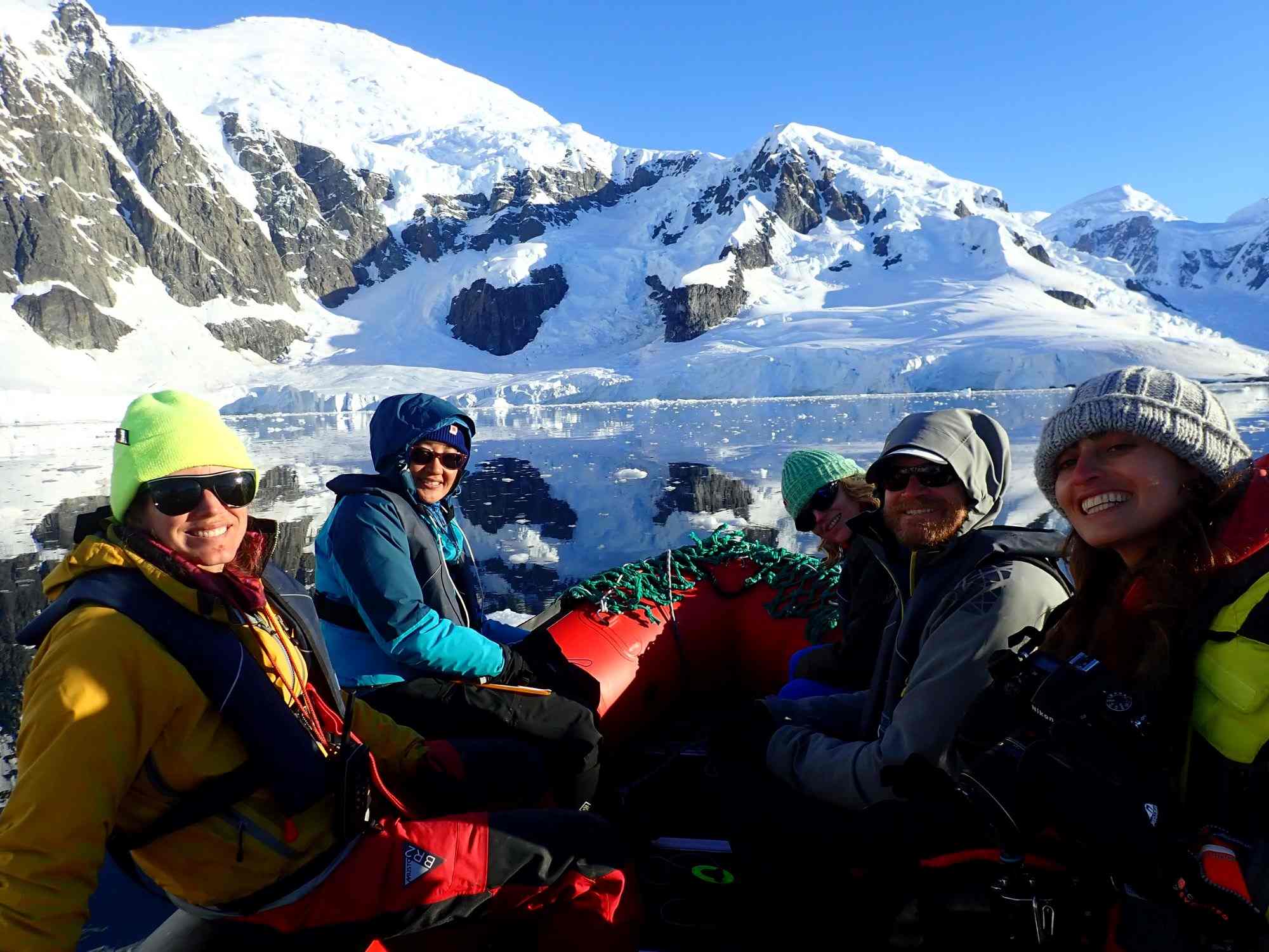 Group of people in a small boat in Antarctic waters, wearing winter gear, with snow-covered mountains in background.