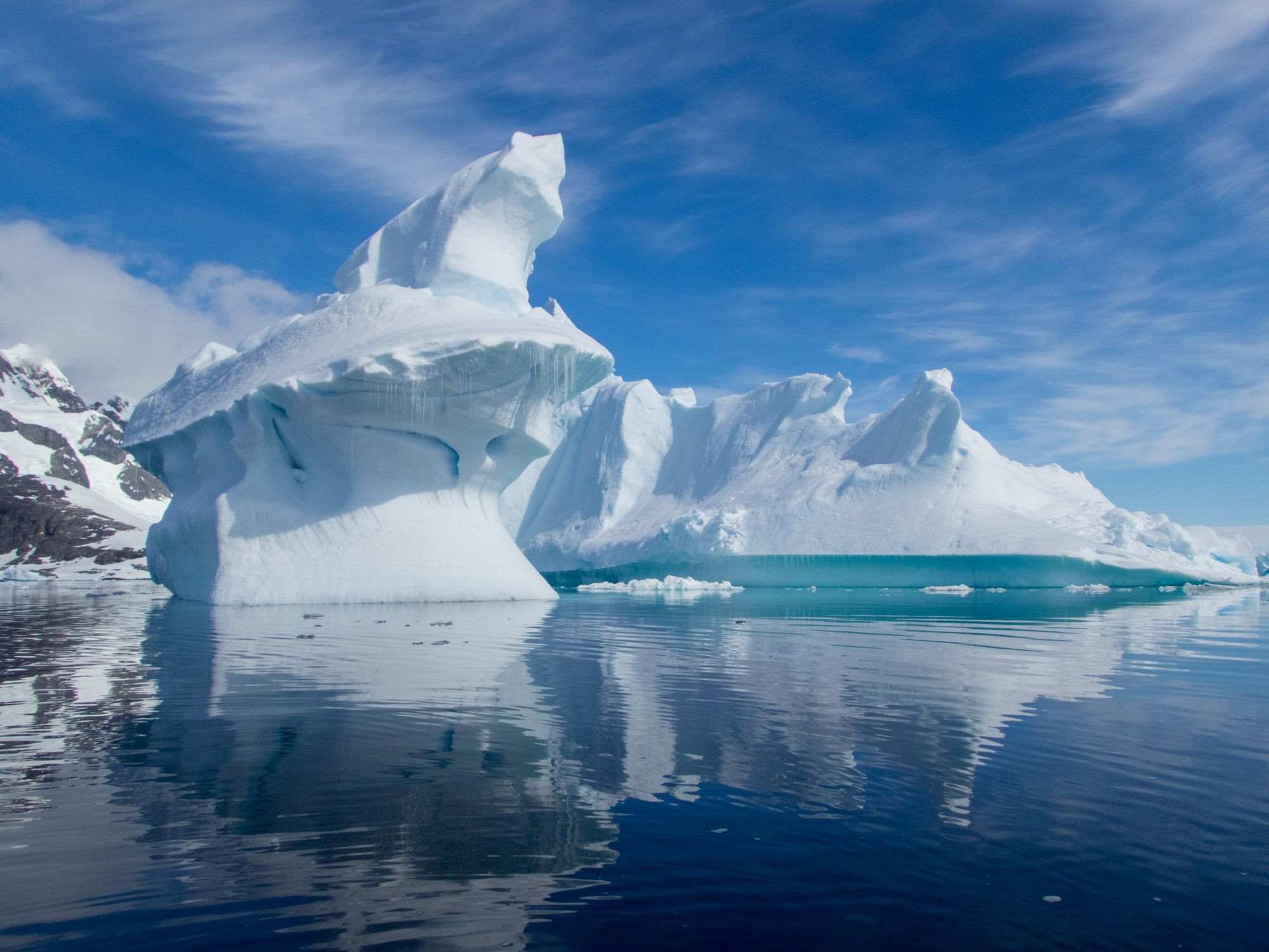 Large iceberg with clear reflection on calm Antarctic waters under a partly cloudy sky, highlighting natural beauty.