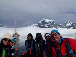 Group of people in winter clothing on boat near snowy mountains and calm water in Antarctica during cloudy weather.