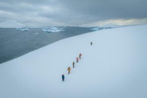 Group of people hiking on snowy terrain in Antarctica with icebergs and snow-covered mountains in the background under a cloudy sky.