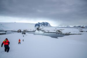 People in colorful winter gear walking towards a body of water in Antarctica, with snow-covered mountains and a cloudy sky in the background.