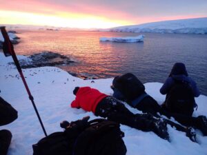 Group of people lying on snow near icy water with floating icebergs and a warm sunrise/sunset glow in Antarctica.