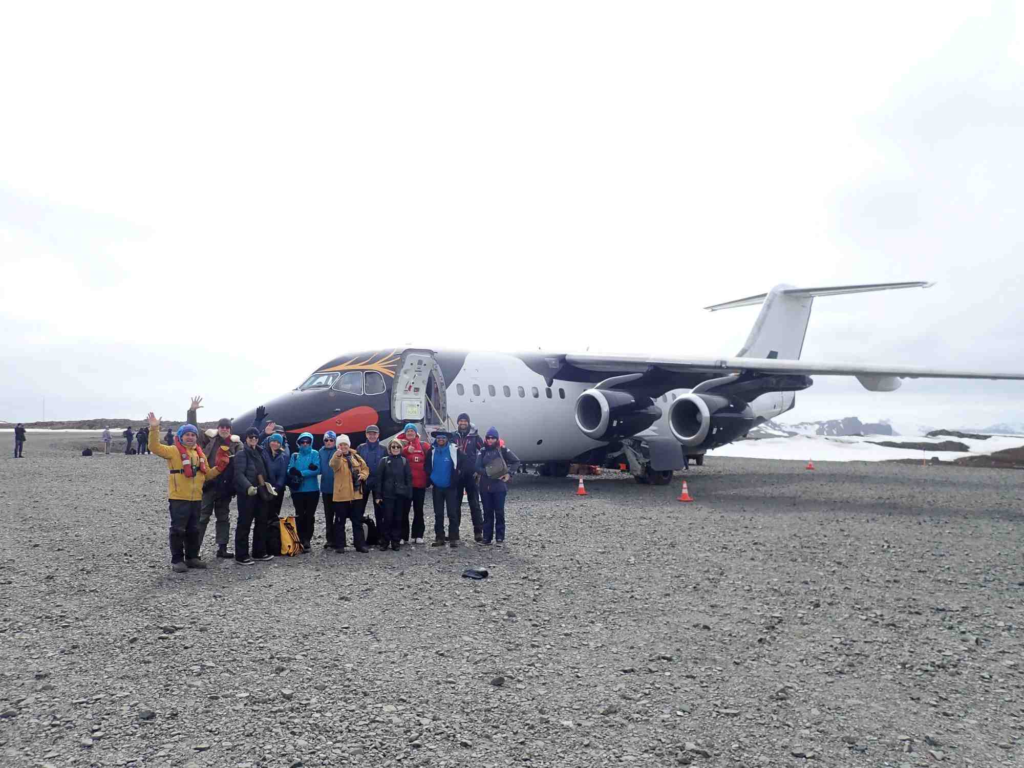 Group of people standing in front of a four-engine airplane on a gravel surface, dressed in outdoor gear, with overcast sky in Antarctica.