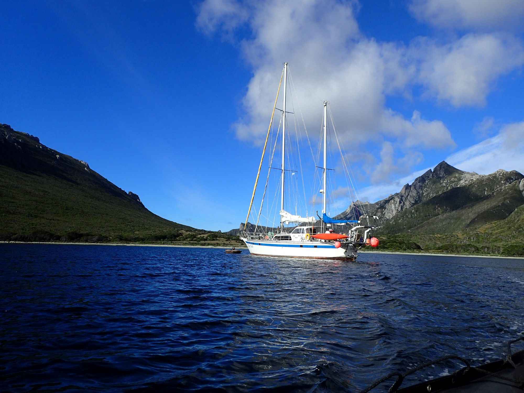 Sailboat anchored near a mountainous shore with green vegetation and rocky terrain, partly cloudy sky, Antarctica.