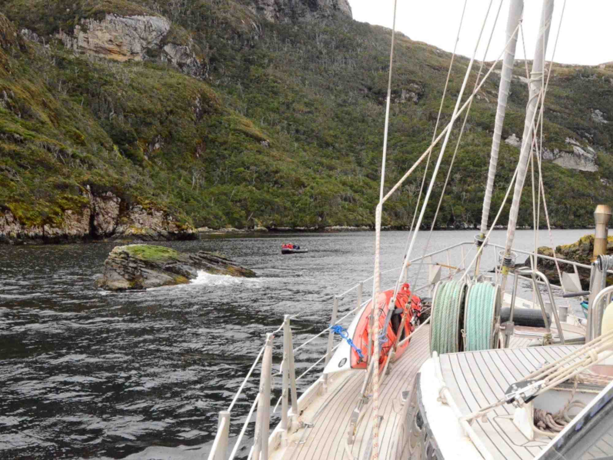 Sailboat docked near a rocky shore with green hillside, calm waters, and inflatable boat in the distance, exploration setting.