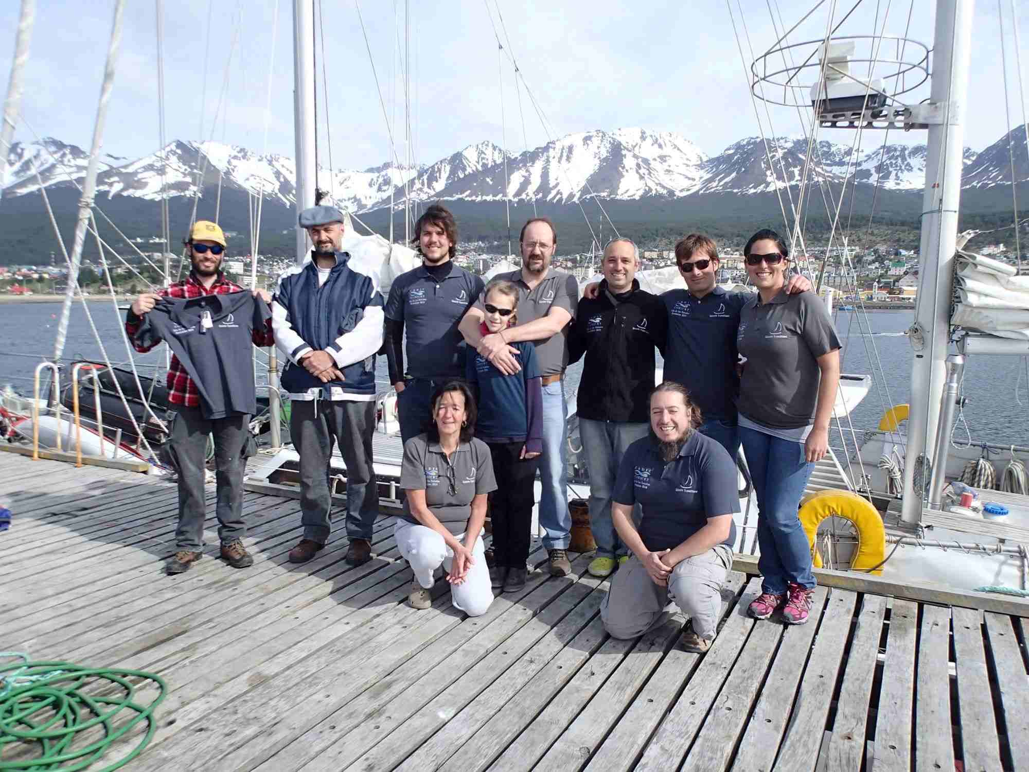 Group on a dock with a sailboat, snow-capped mountains, and blue sky in the background, Antarctica.