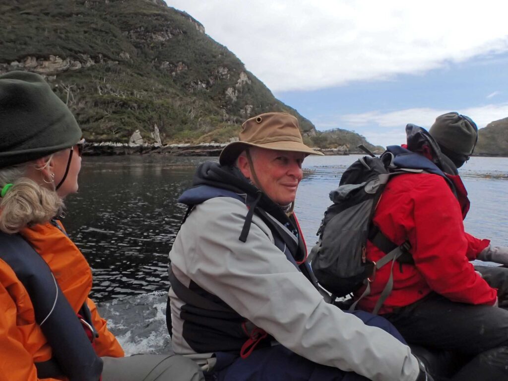 Three people in jackets and hats sitting in a boat on calm Antarctic waters, with rocky, vegetated shoreline in the background.