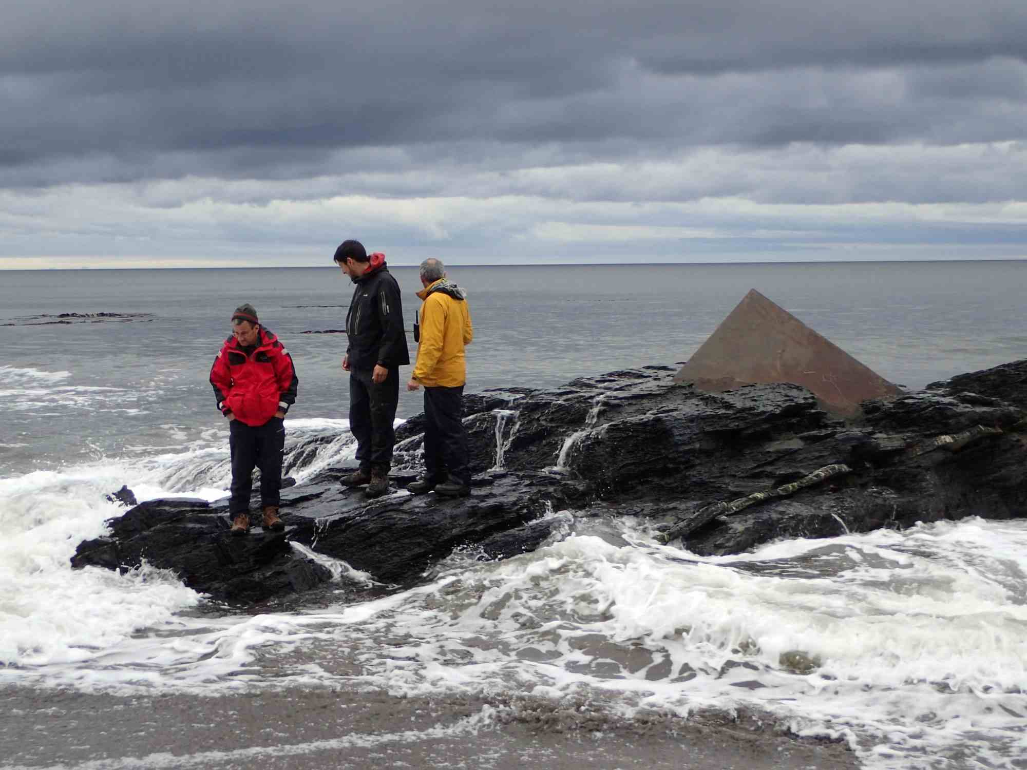 Three people in colorful jackets standing on rocky shore with rough sea and overcast sky in Antarctica, waves crashing around them.