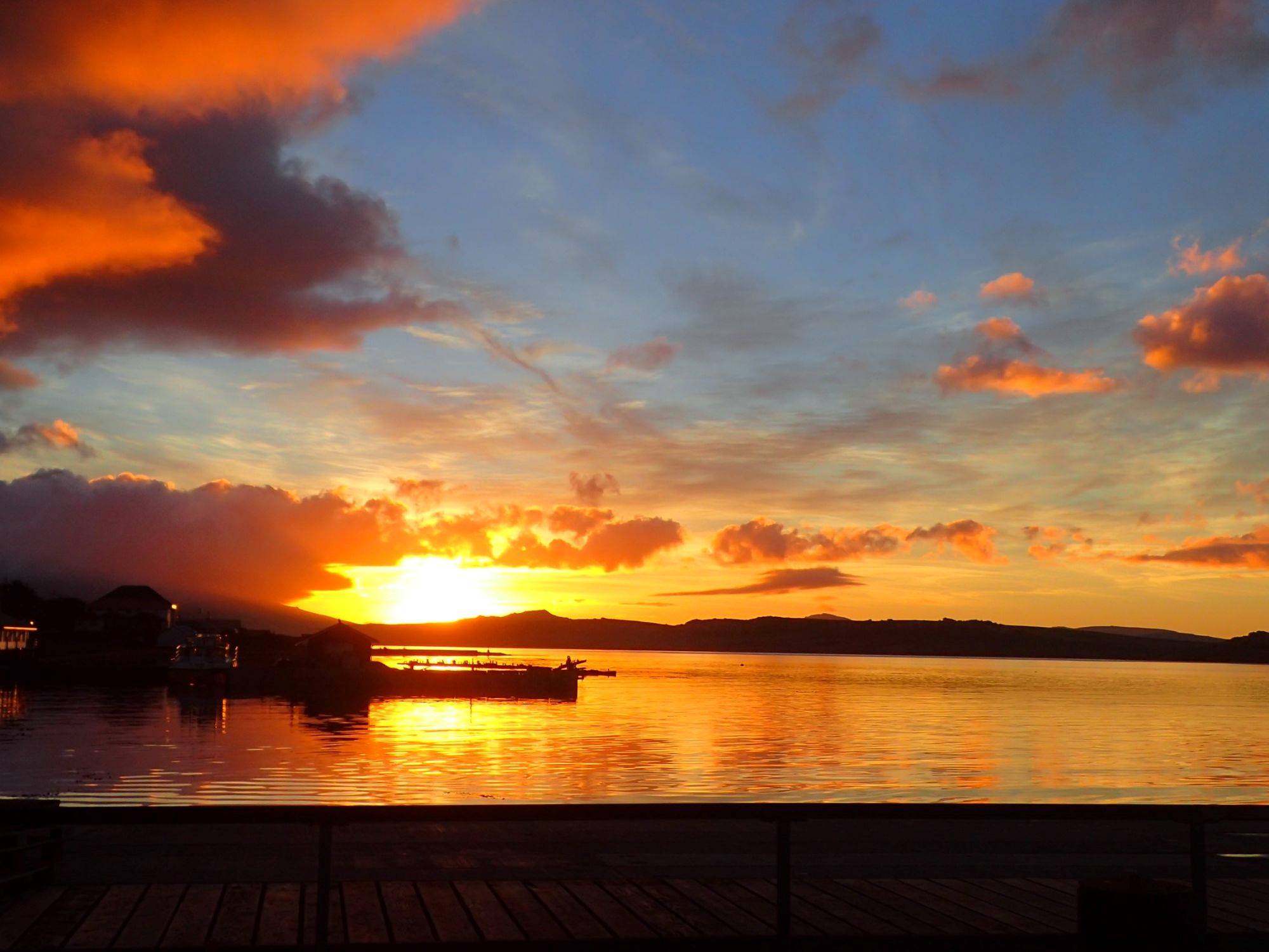 Sunset over Antarctic waters with a dock in the foreground, vibrant sky colors, and silhouettes of buildings.