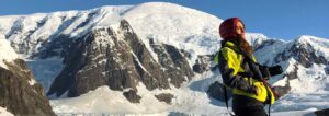 Person in yellow and black jacket with a red hat standing in front of snow-covered mountains in Antarctica, holding a camera.