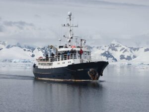 Expedition ship navigating icy Antarctic waters with snow-covered mountains in the background.